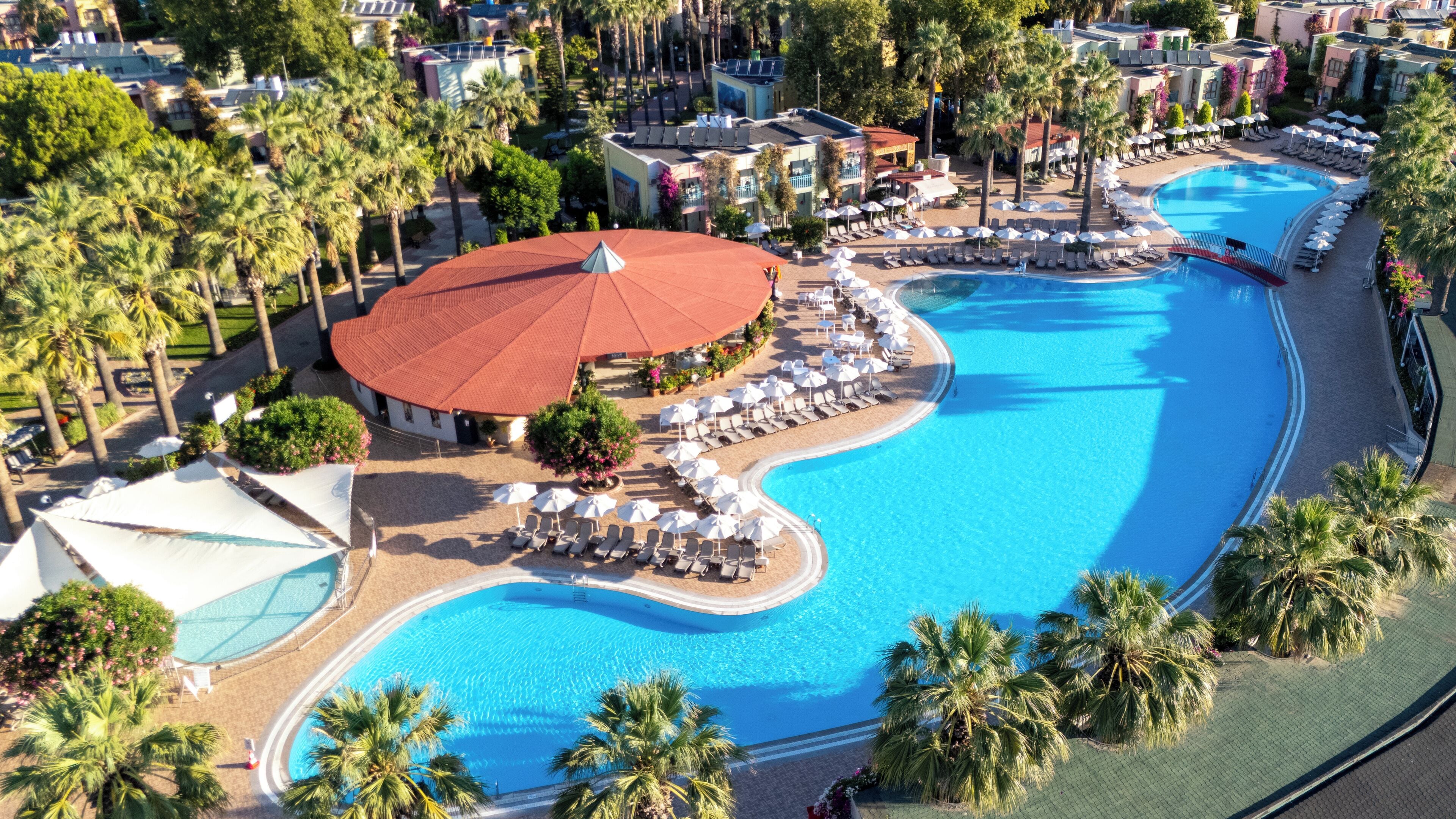 Ein großer Swimmingpool mit Liegestühlen und Sonnenschirmen, umgeben von Palmen und Hotelgebäuden.