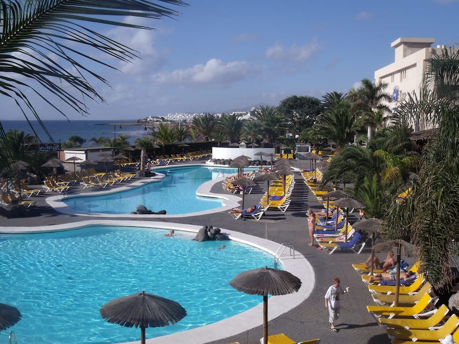 Zona de piscina con múltiples tumbonas y sombrillas, rodeada de palmeras y vistas al océano.