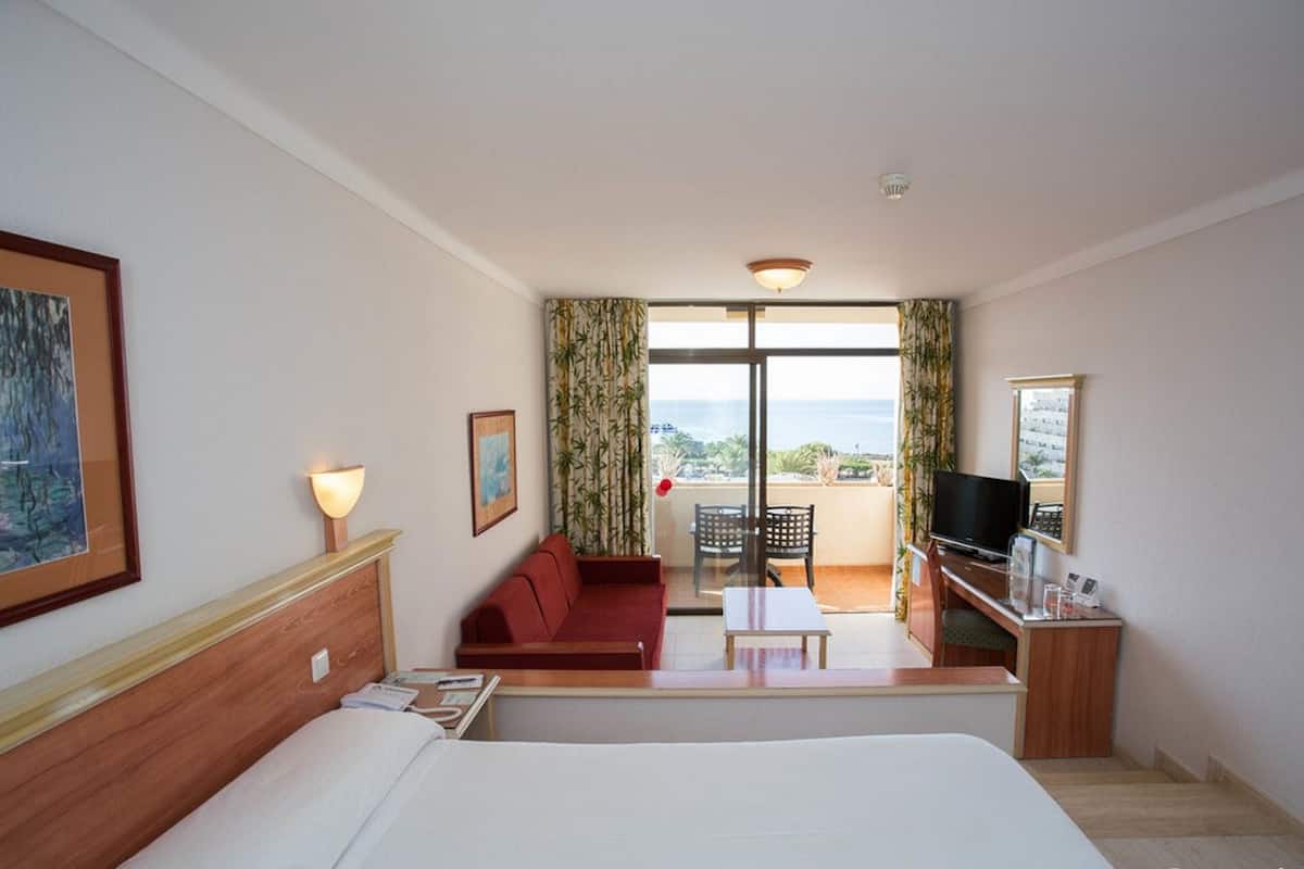 A hotel room with a bed, a red sofa, a small table, a TV, and a balcony with a view.