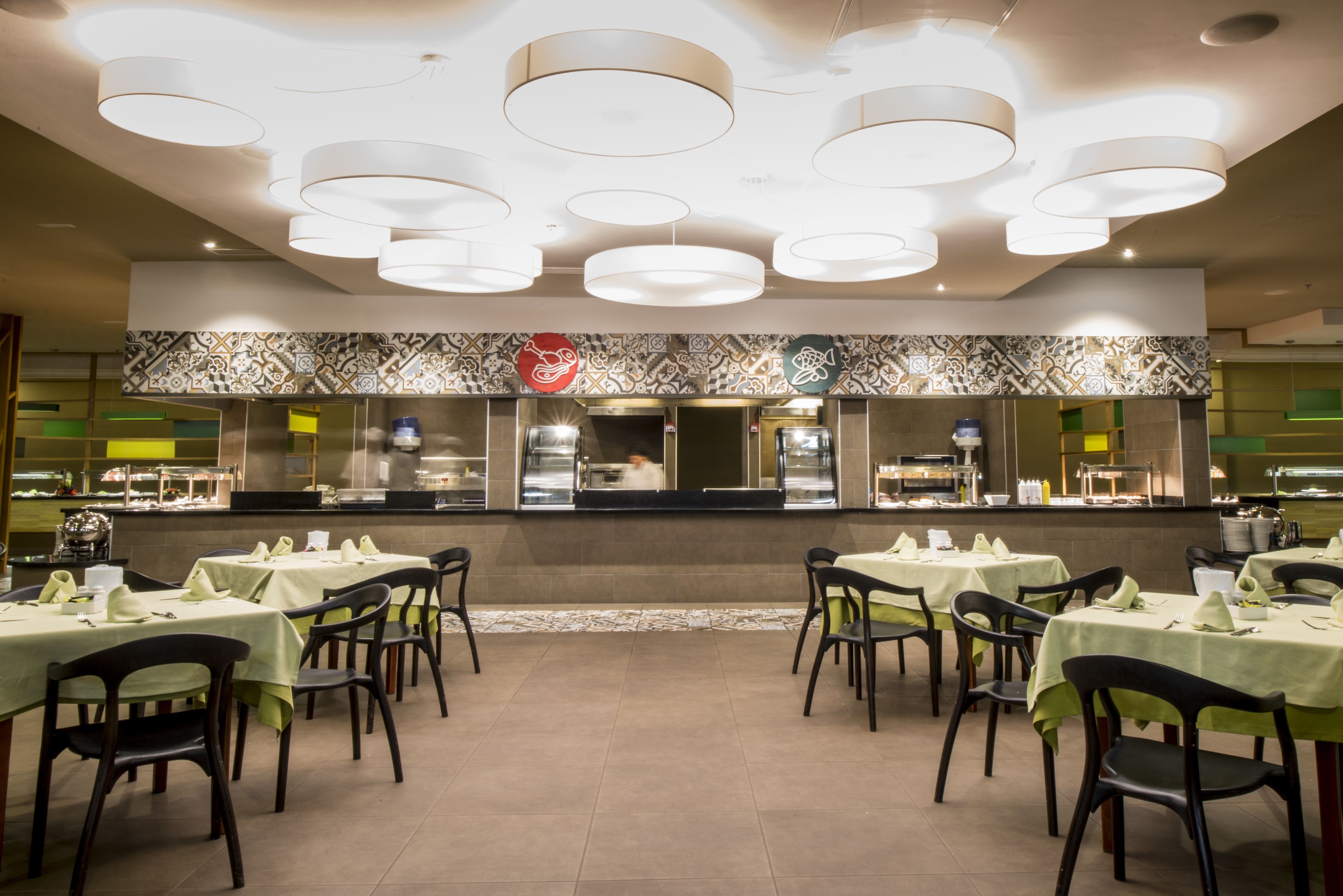 Um restaurante com várias mesas e cadeiras, um balcão com um cozinheiro e uma parede com padrão.