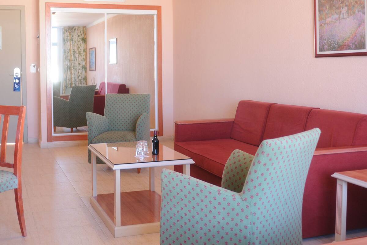A living room with a red sofa, patterned armchairs, a glass coffee table, and a small table with a telephone.