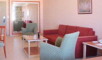 Un salon avec un canapé rouge, des fauteuils à motifs, une table basse en verre et une petite table sur laquelle se trouvent un téléphone et des papiers.
