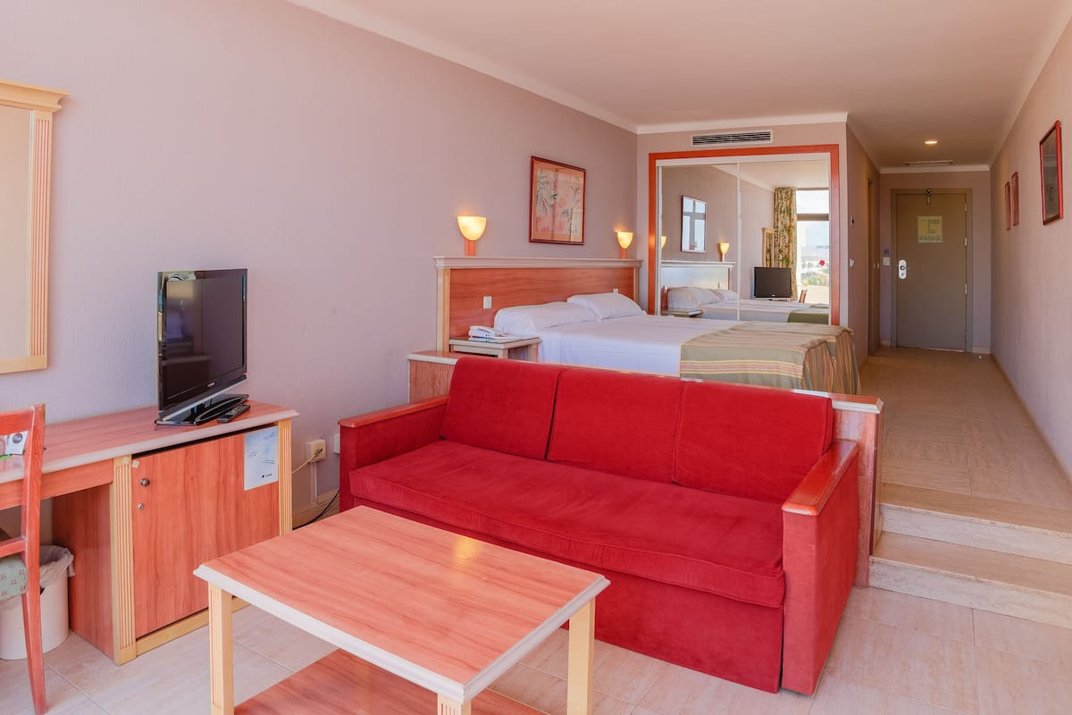 A hotel room with a bed, a red sofa, a wooden desk with a TV, and a small table.