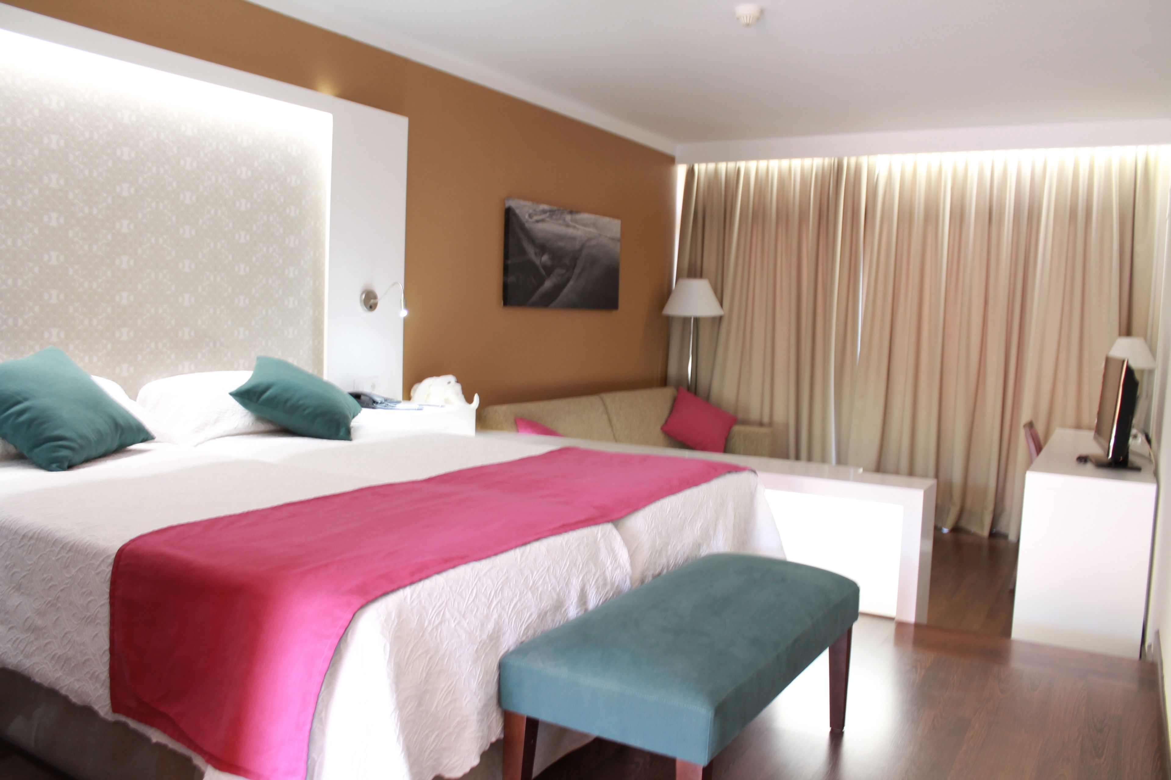 A hotel room with a bed, a desk, a bench, a lamp, and a painting on the wall.
