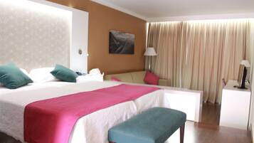 A hotel room with a bed, a desk, a bench, a lamp, and a painting on the wall.