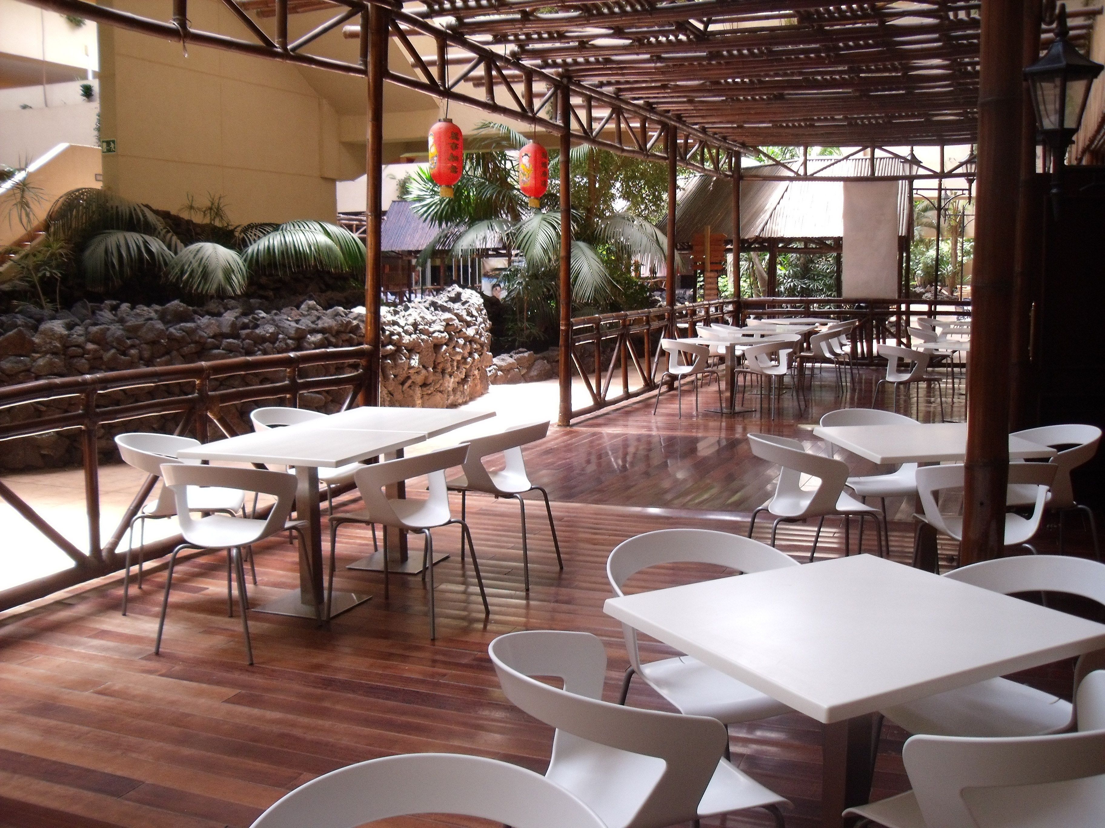 Un espacio exterior para comer con mesas y sillas blancas, estructuras de bambú y un muro de piedra con plantas.