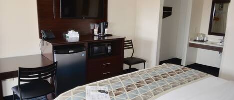 Una habitación de hotel con una cama, un escritorio con teléfono y microondas, una silla y un televisor en la pared.