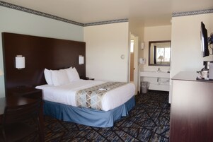 A hotel room with a large bed, a desk, a chair, a dresser, a mirror, and a bathroom with a sink and toilet.