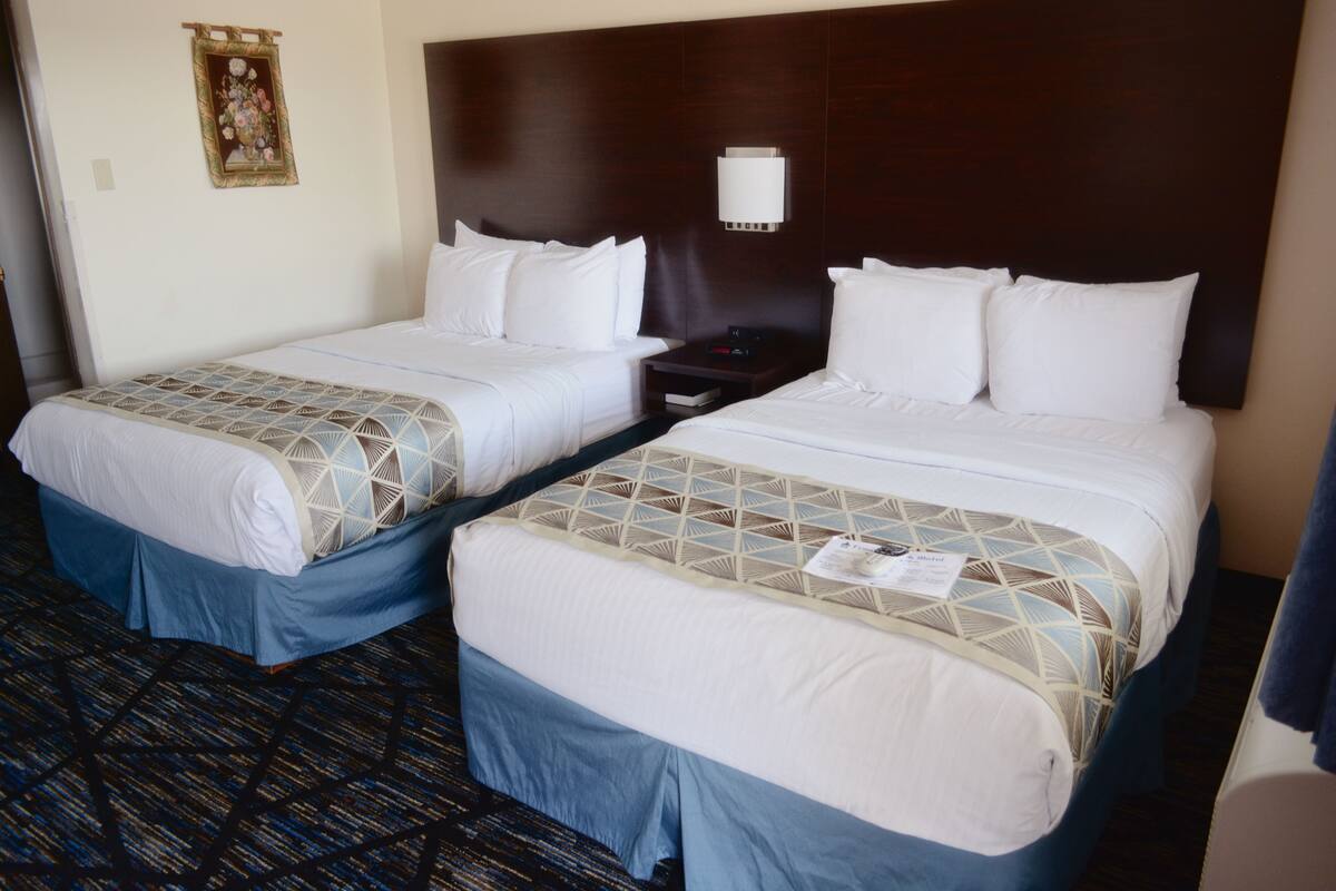 A hotel room with two beds, each with a patterned bedspread and blue skirts. There is a nightstand with a lamp between the beds, and a wall-mounted light above the headboard.