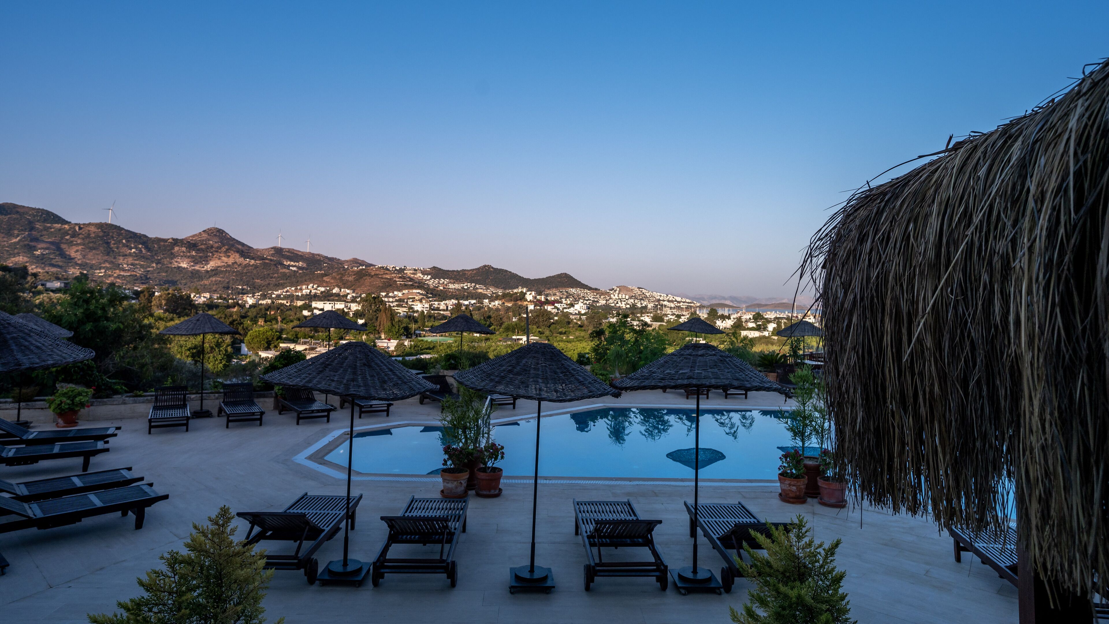 A poolside area with lounge chairs and umbrellas, a pool, and a view of a town and mountains in the background.
