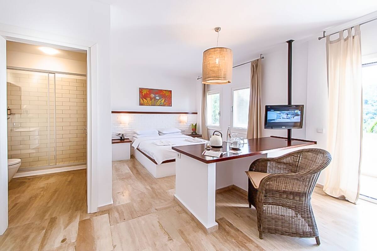 Quarto de hotel com cama, escrivaninha com cadeira, televisão e banheiro com chuveiro.