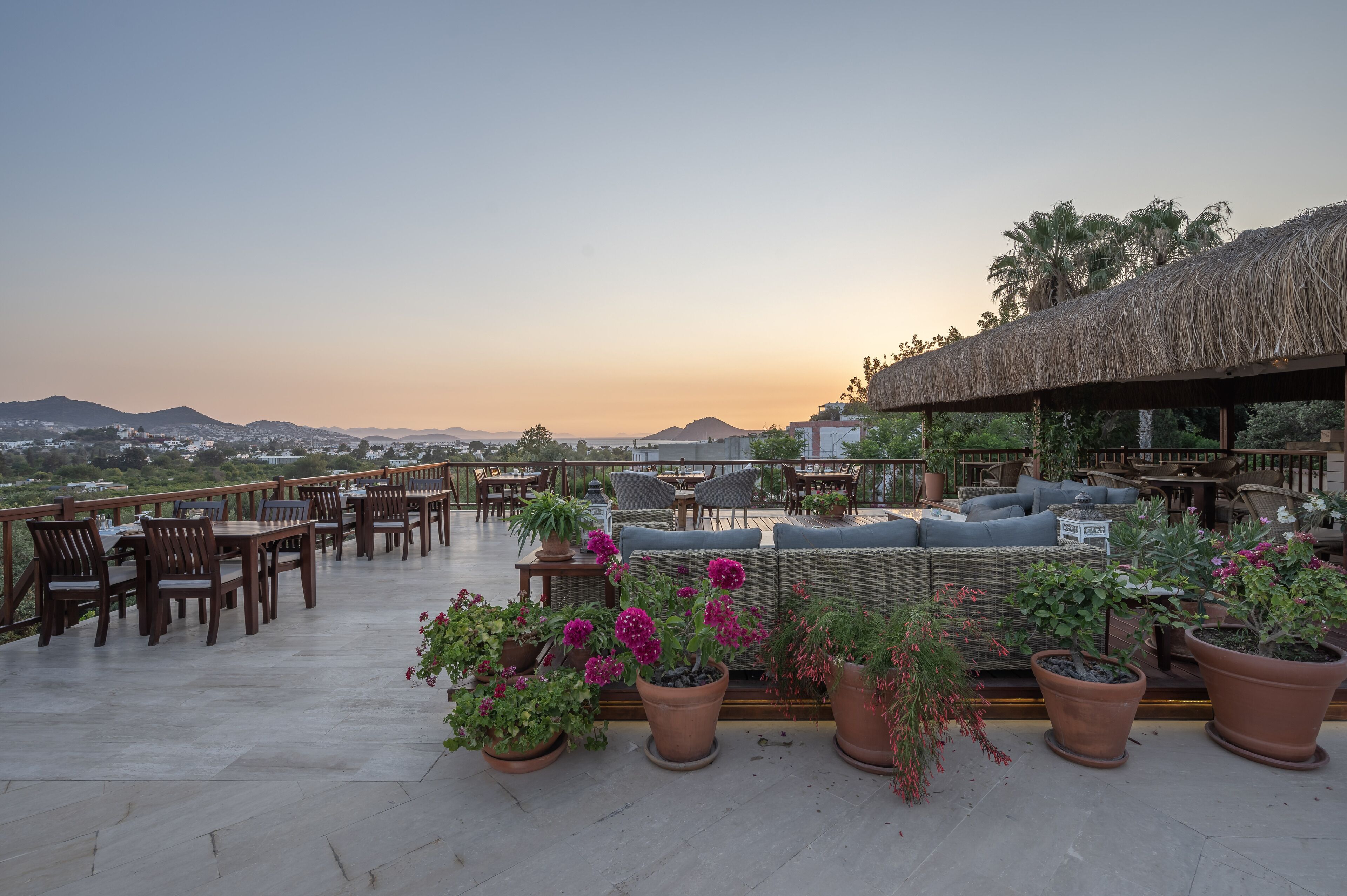 Una terraza en la azotea con mesas y sillas de madera, plantas en macetas y una zona de estar con techo de paja.