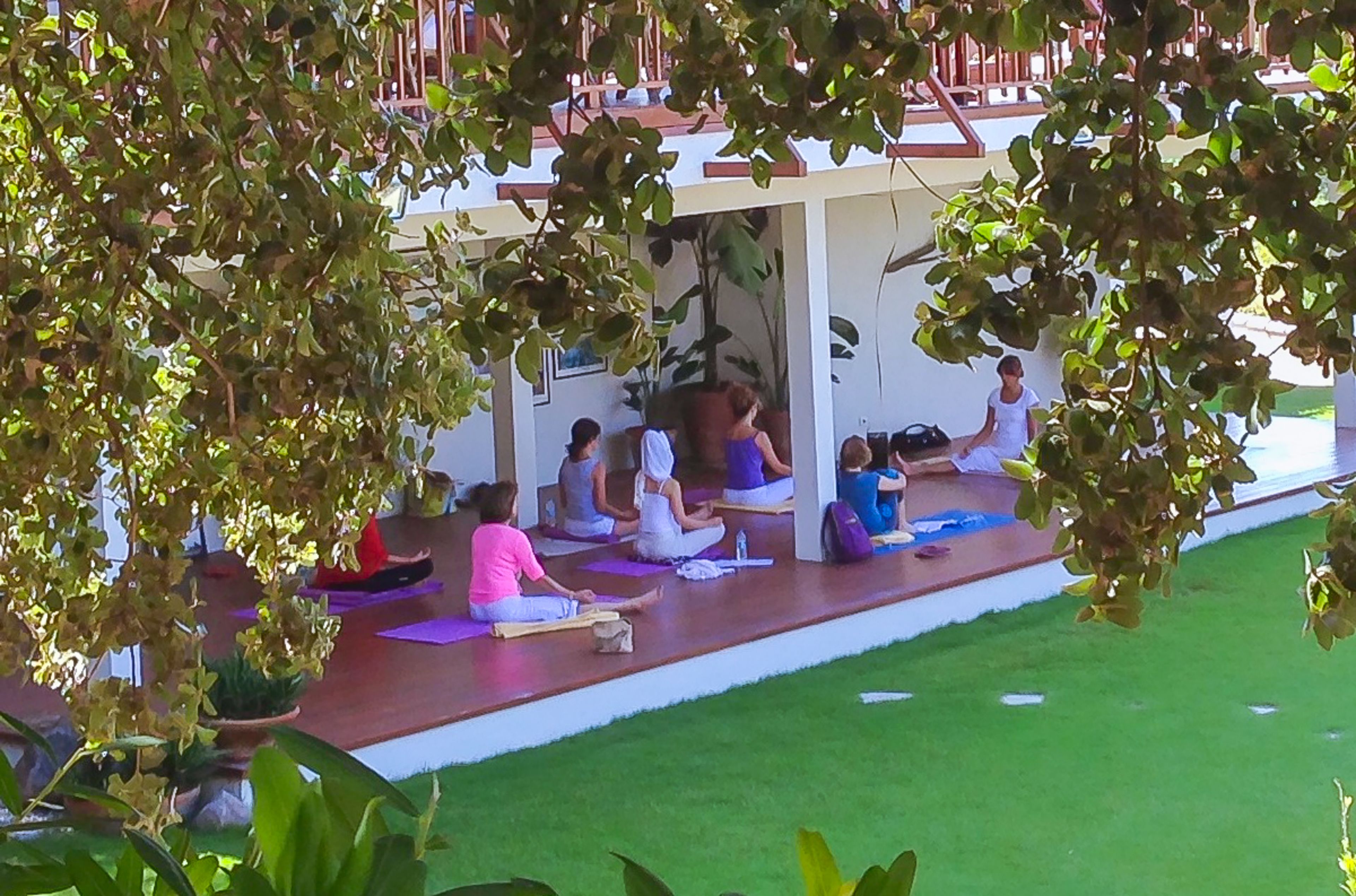 Um grupo de pessoas sentadas em tapetes de yoga em área externa coberta, com parede branca e deck de madeira.
