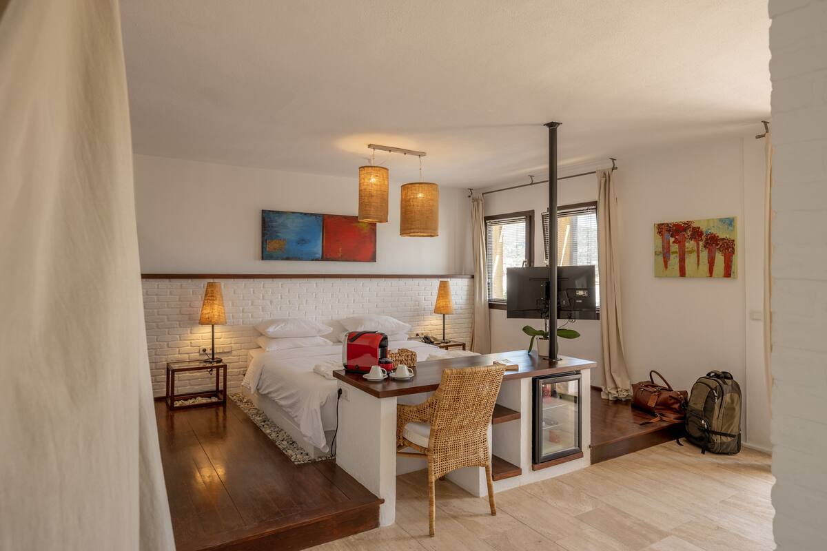 Quarto de hotel pequeno com cama, abajures, uma secretária com cadeira, televisão e um quadro na parede.