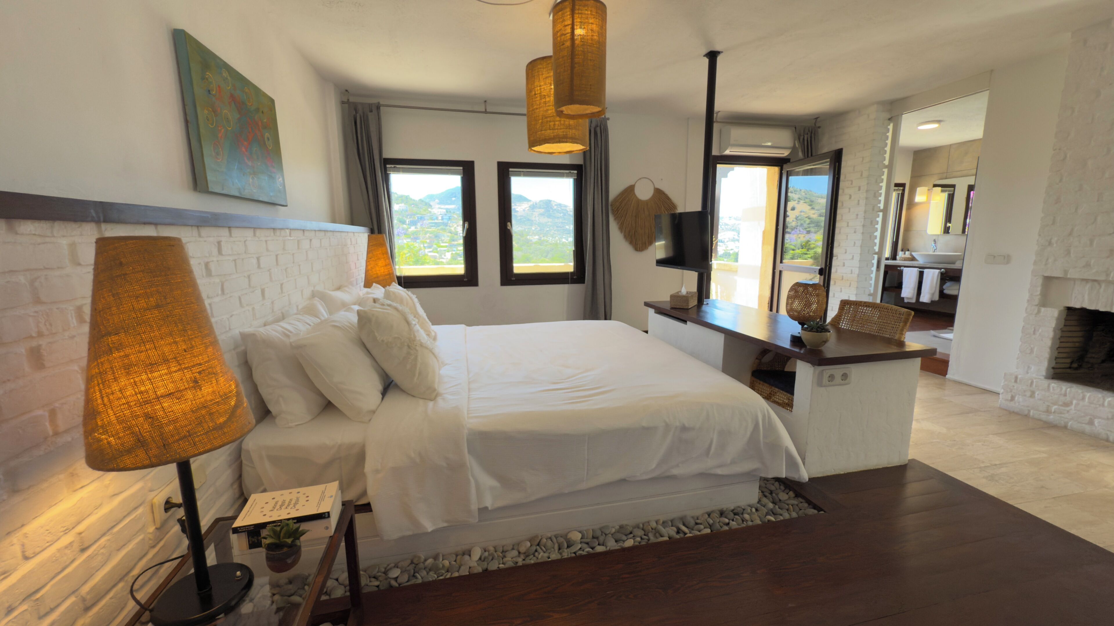A modern bedroom with a large bed, a desk, a fireplace, and a view of the outdoors.