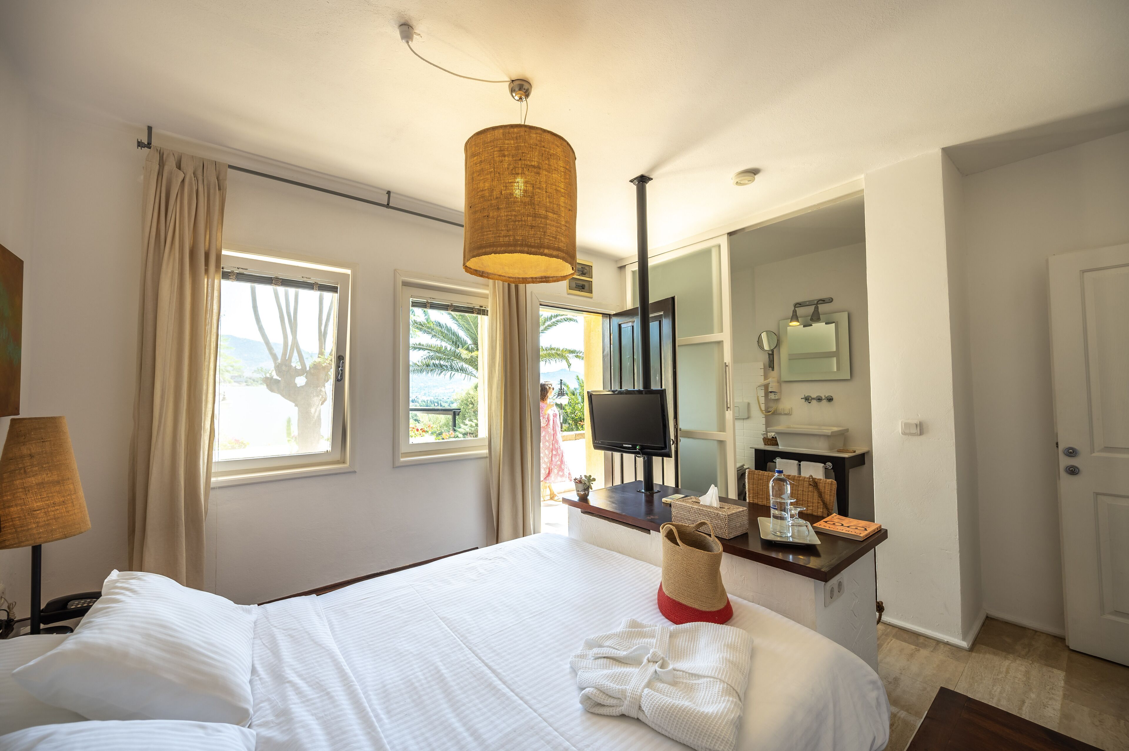Quarto de hotel com cama, televisão, espelho, banheiro e janela com cortinas.