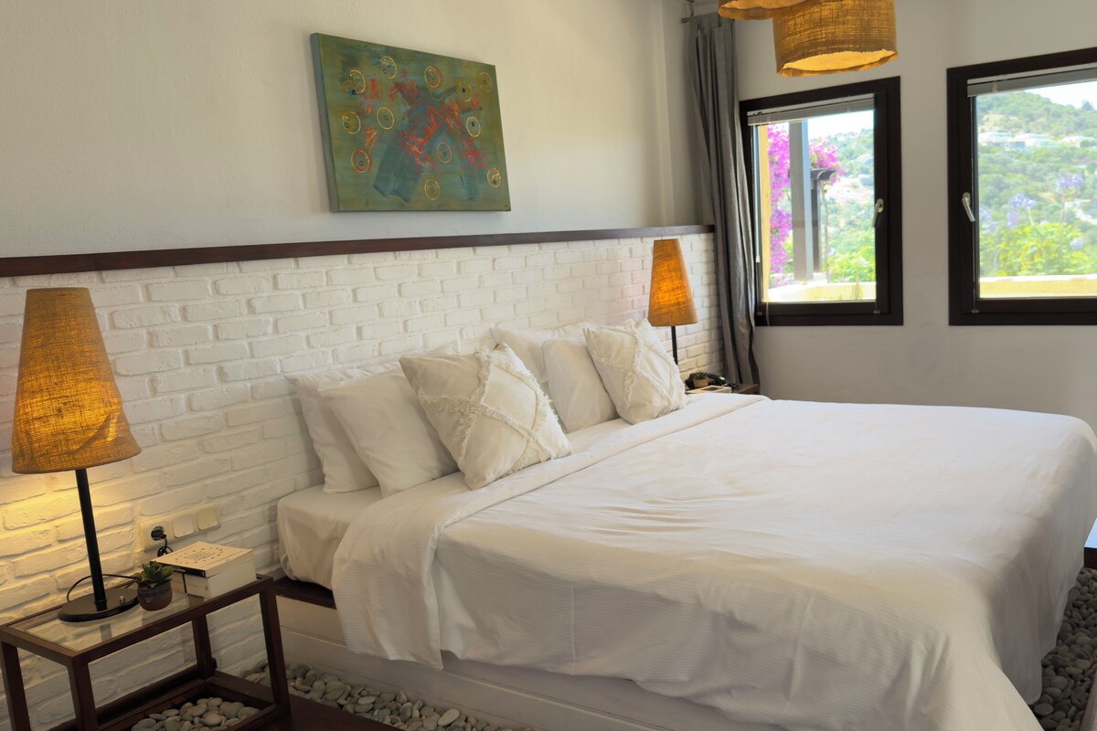 Una cama bien tendida con sábanas blancas, un cabecero con diseño de ladrillo, un cuadro colgado en la pared y una ventana con vistas a la vegetación.