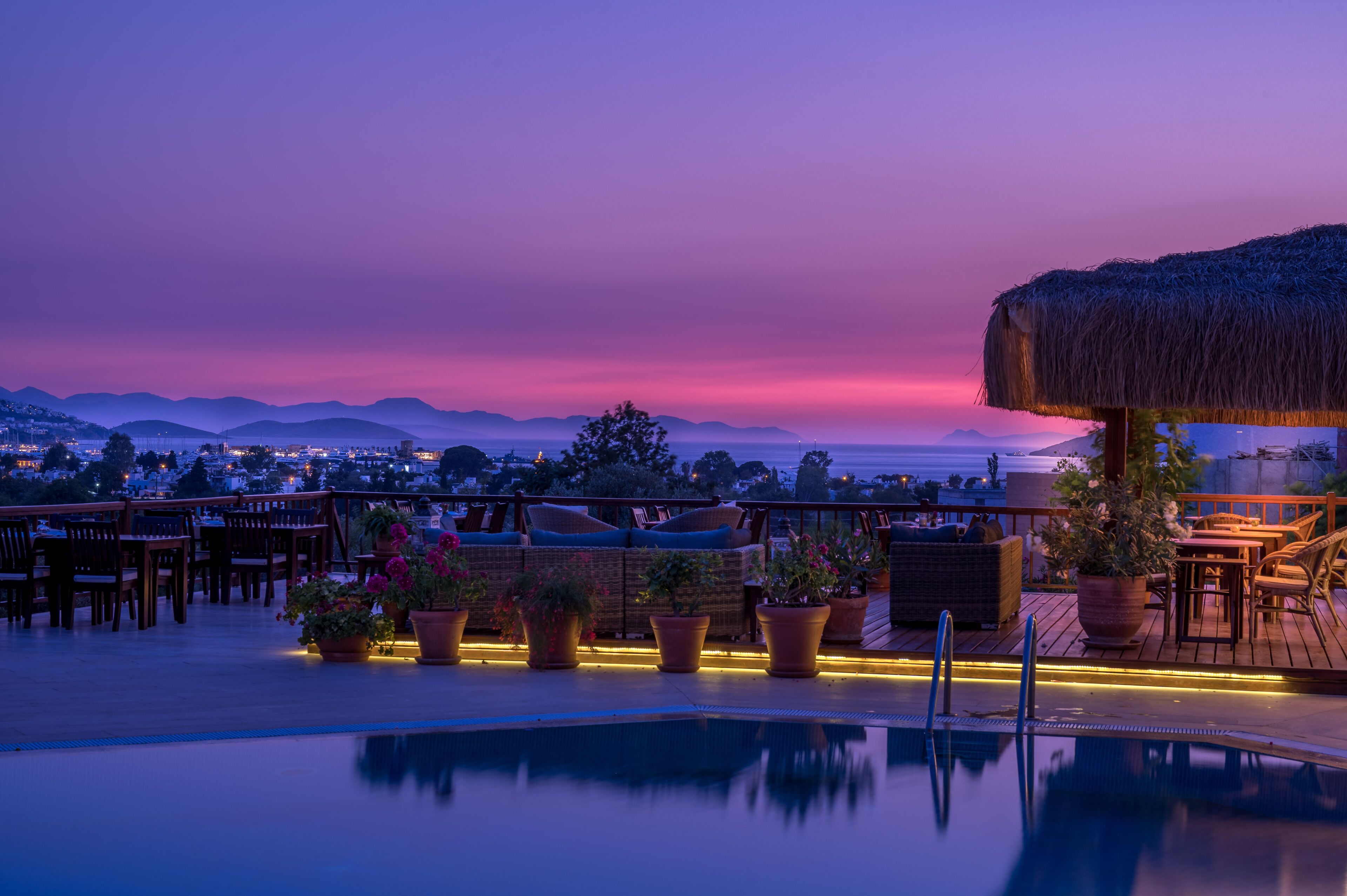 A rooftop pool area with lounge chairs and potted plants, overlooking a cityscape and mountains at dusk.