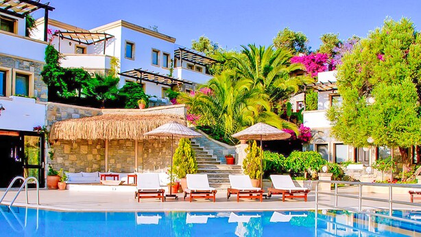 A swimming pool with lounge chairs and umbrellas, surrounded by lush greenery and a multi-story building. - 4Reasons Hotel And Bistro | 12+ (Bodrum)