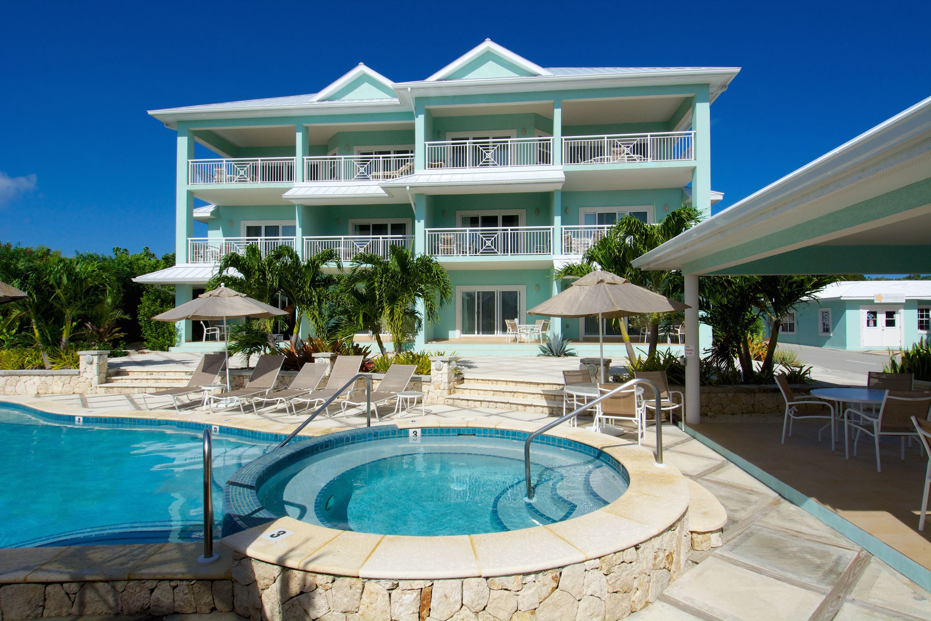 Un resort con piscina, sillas de descanso, sombrillas y un edificio de varios pisos con balcones.