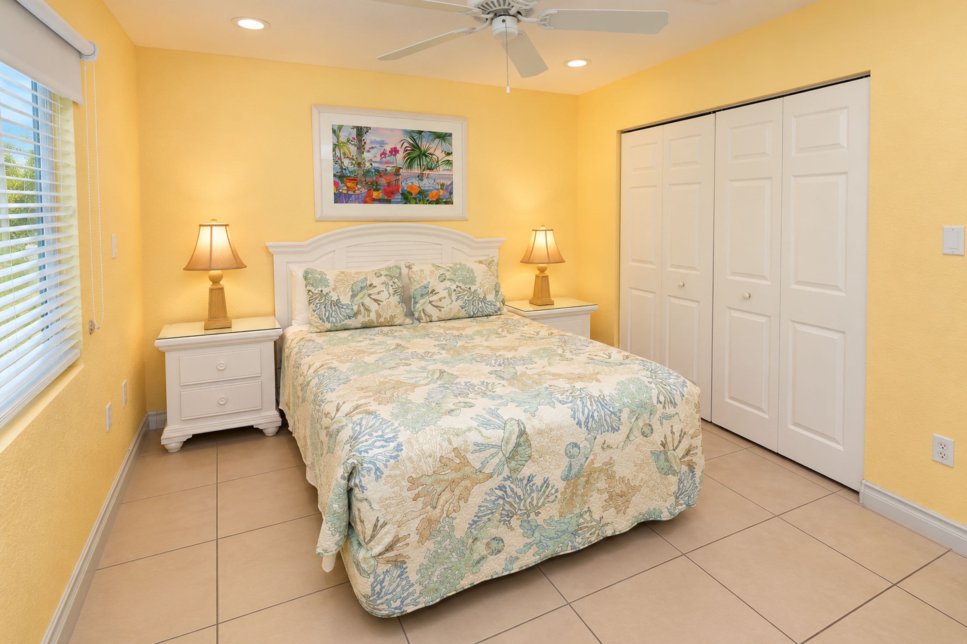 A bedroom with a bed, two bedside tables with lamps, a ceiling fan, a door, and a window with blinds.