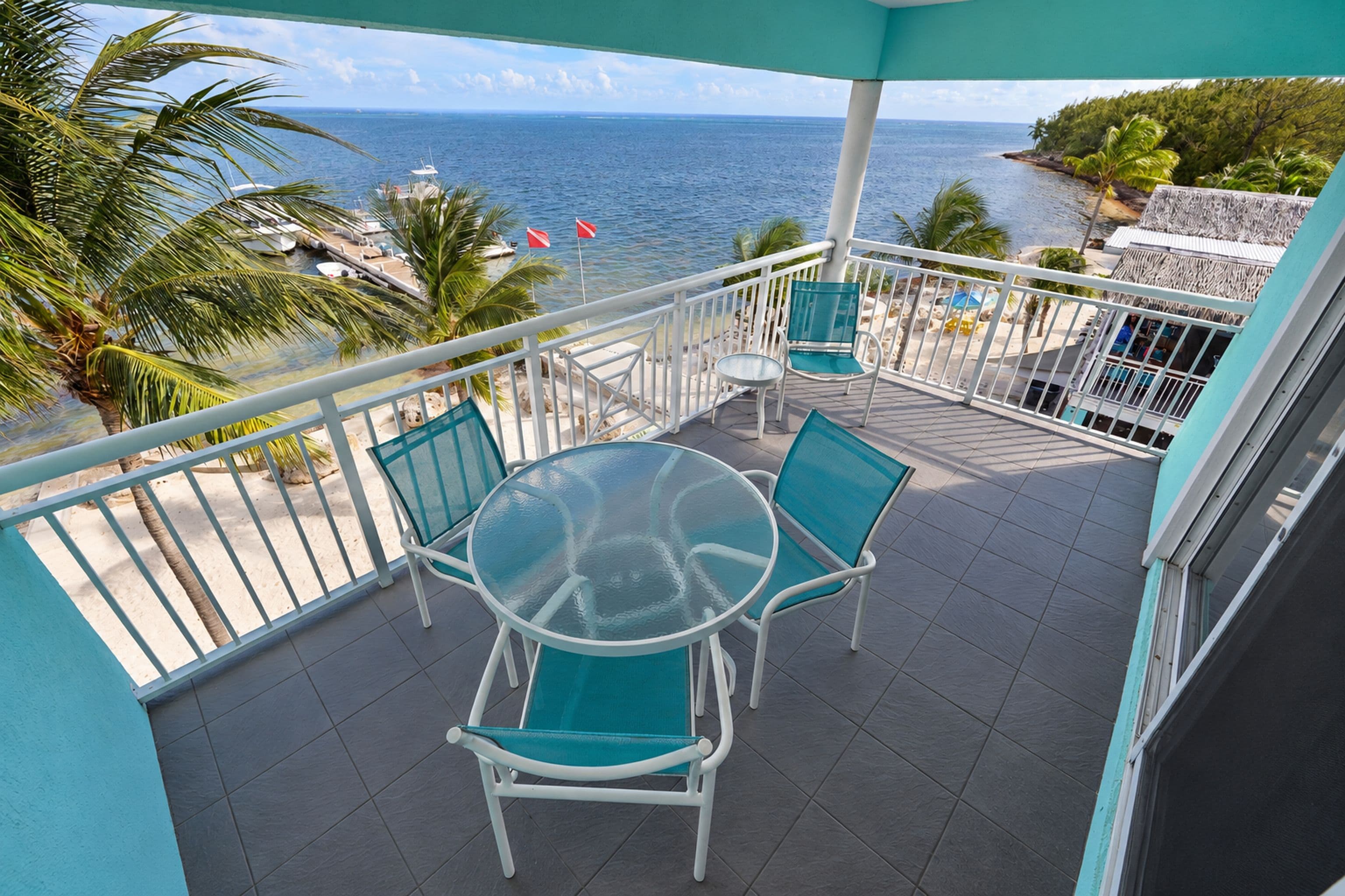 A balcony with a glass table and chairs, overlooking a beach and the sea.