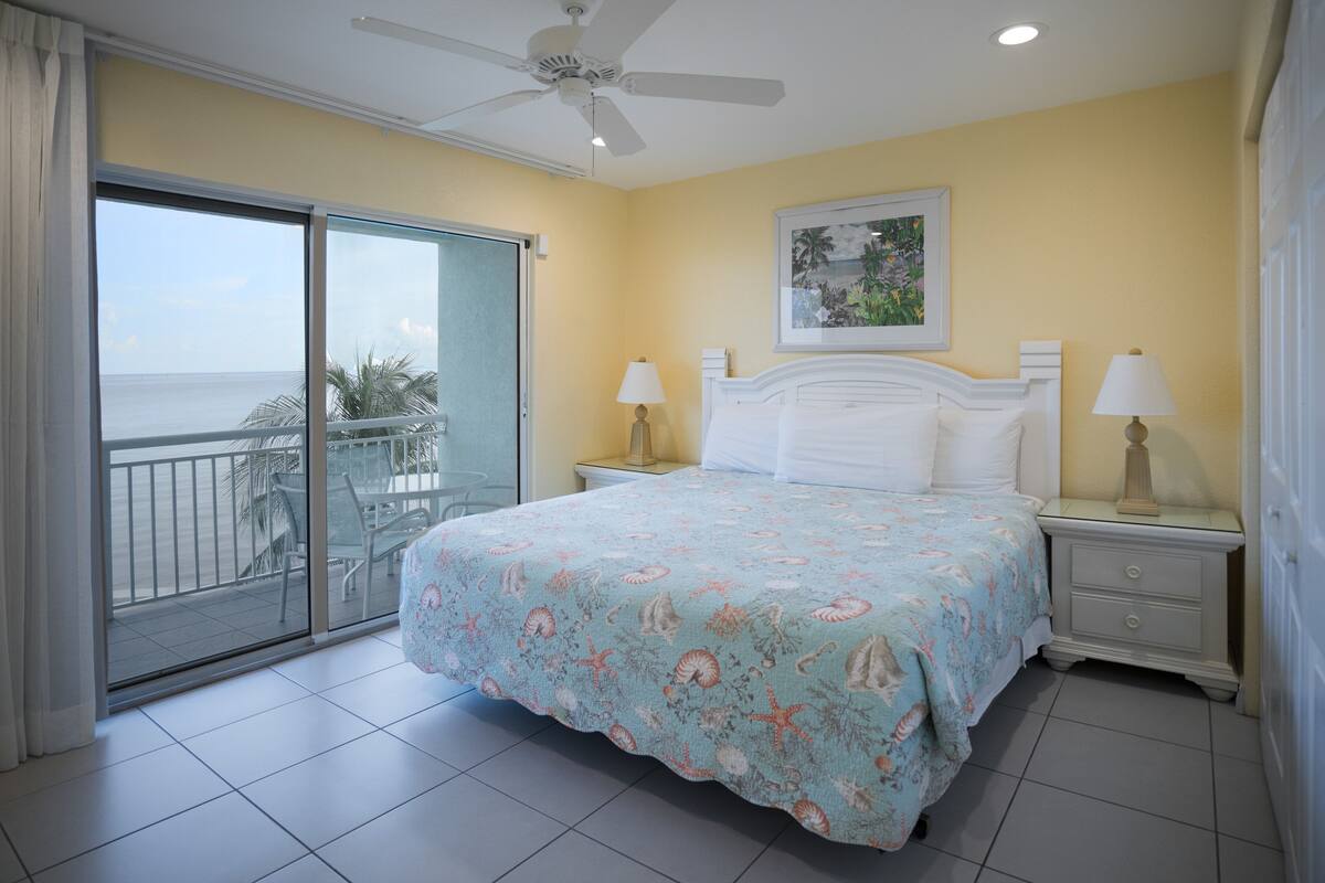 A bedroom with a balcony, a bed with a floral bedspread, two bedside tables with lamps, and a ceiling fan.