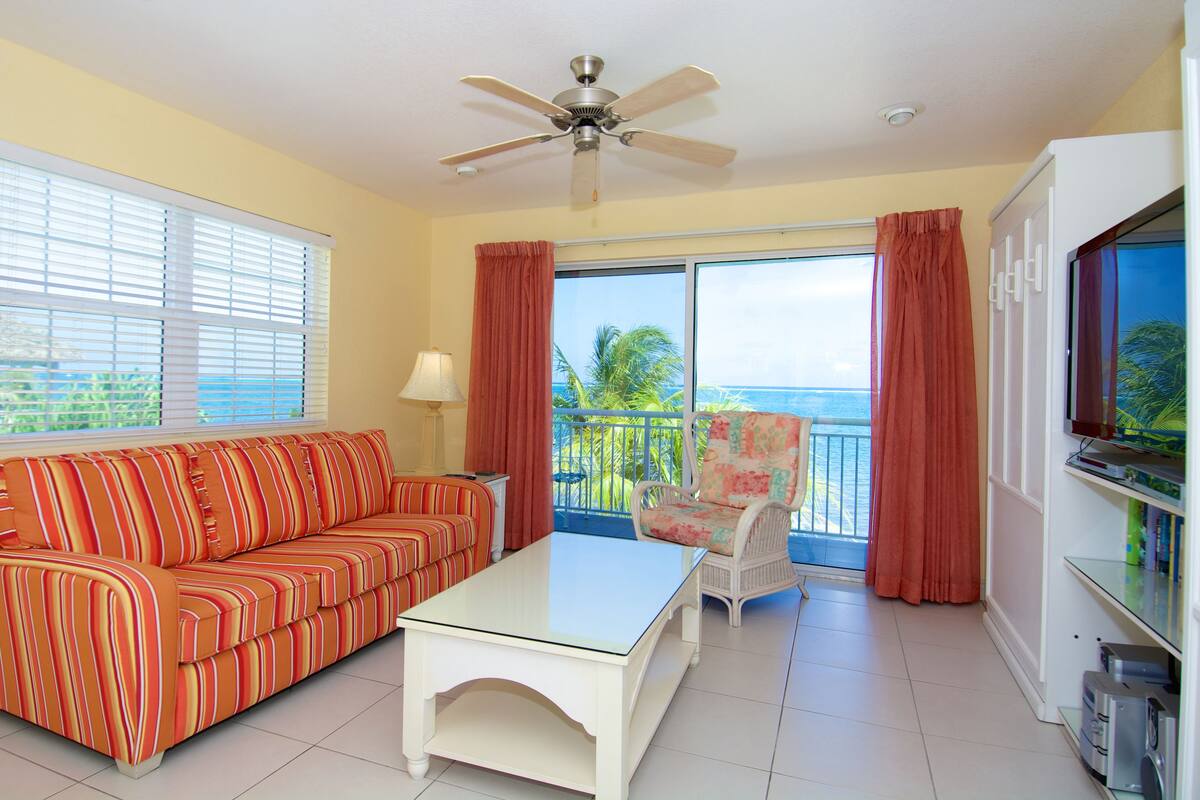 A room with a striped sofa, a glass coffee table, a chair, a television, and a balcony with curtains.
