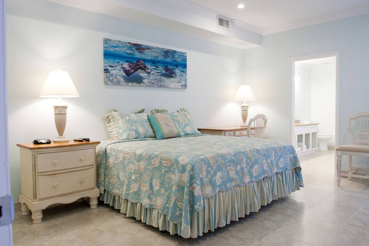 A bedroom with a bed, two bedside tables with lamps, a chair, a painting on the wall, and a bathroom visible through an open door.