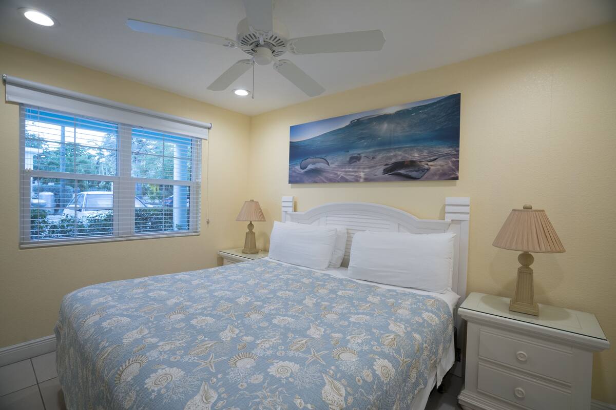A bedroom with a bed, a ceiling fan, a window with blinds, a nightstand with a lamp, and a painting on the wall.