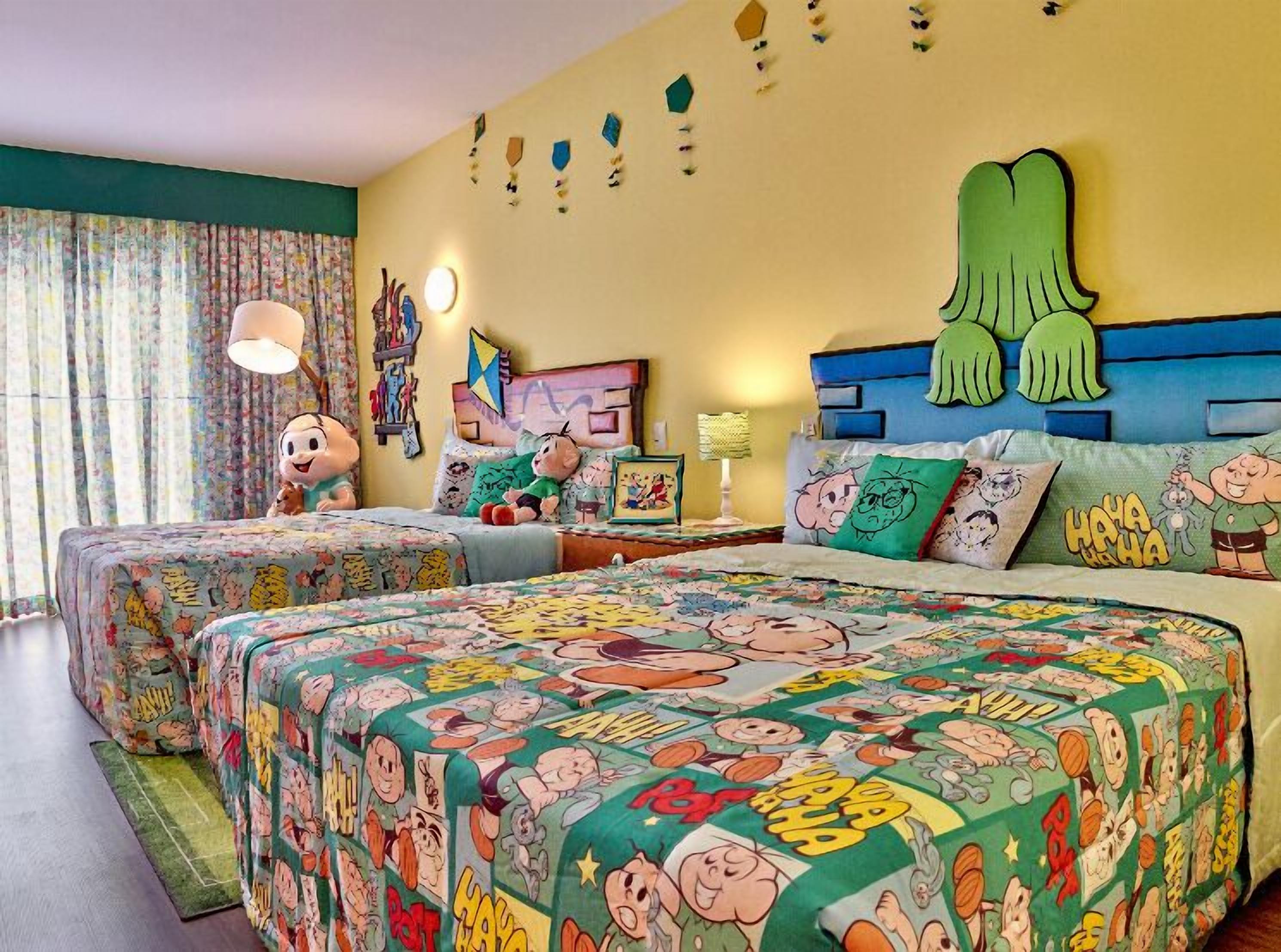 Een hotelkamer met twee bedden, kleurrijk beddengoed met tekenfilmthema, een groot groen kussen en een muur met verschillende decoraties.