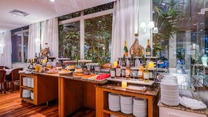 Montaje de bufé con diversos alimentos y bebidas, rodeado de mobiliario de madera y vistas a zonas verdes al exterior.