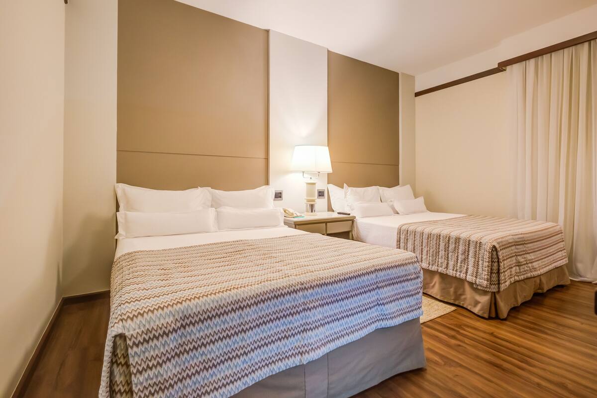 A hotel room with two beds, a nightstand, a lamp, and a window with curtains.