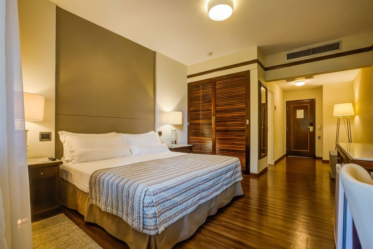 A hotel room with a large bed, bedside lamps, a wooden wardrobe, and a desk with a chair.