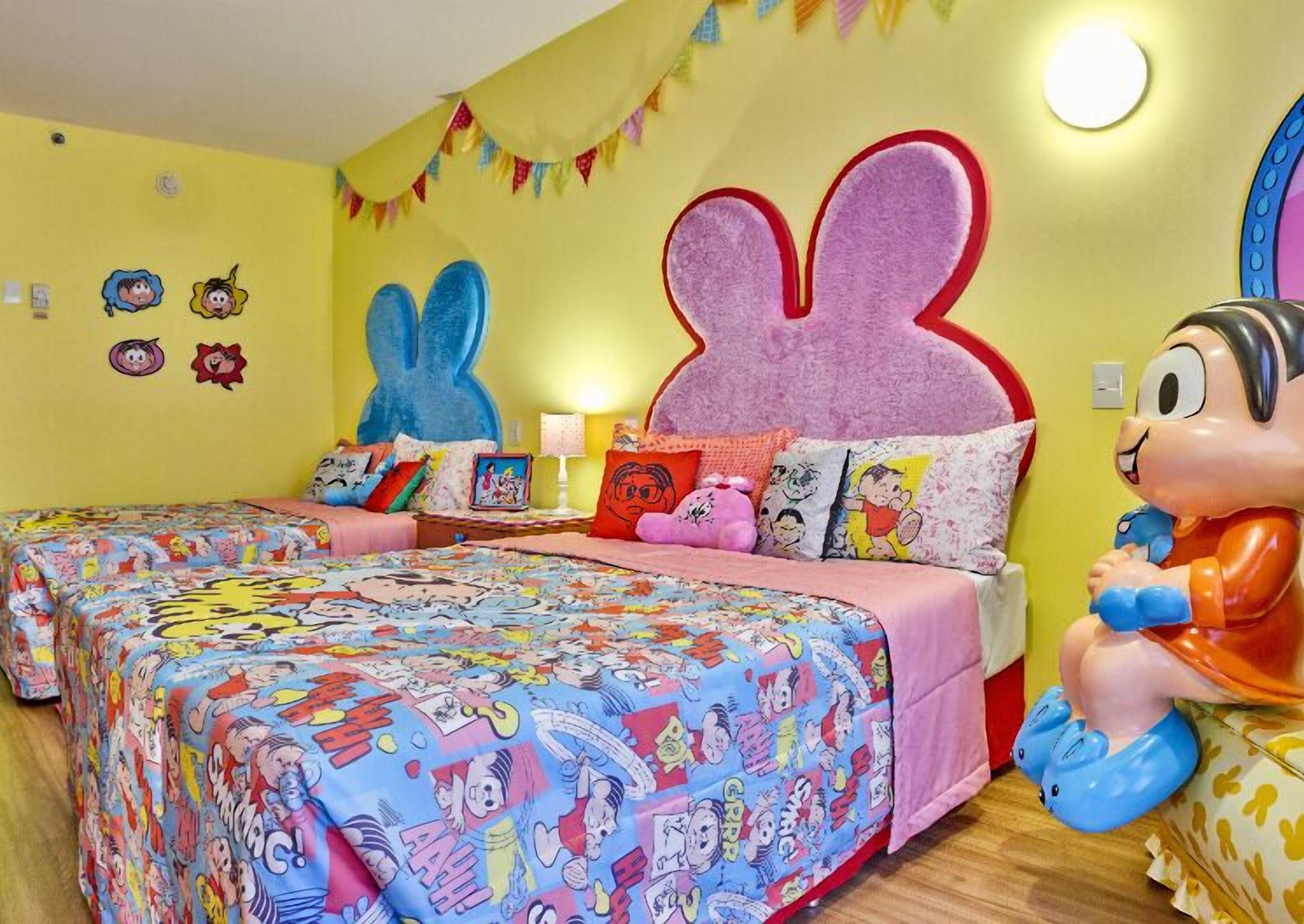 Une pièce aux couleurs vives, avec deux lits, chacun recouvert d’une couverture aux motifs colorés inspirés de bandes dessinées. Les murs sont décorés de personnages de dessins animés et une grande tête de lapin orne le haut du lit.