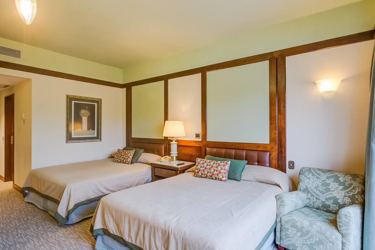 A hotel room with two beds, a nightstand, a lamp, a chair, and a framed picture on the wall.