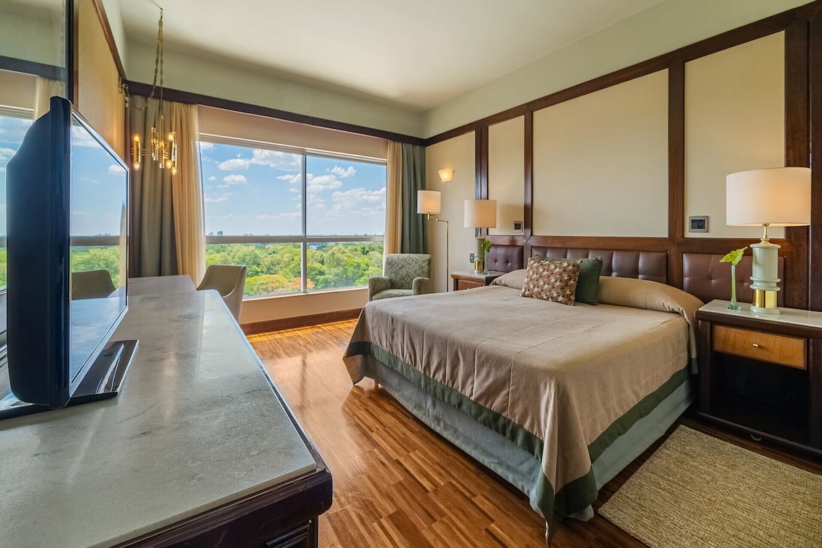 A hotel room with a large bed, a desk, a chair, and a TV. There is a window with a view of greenery.