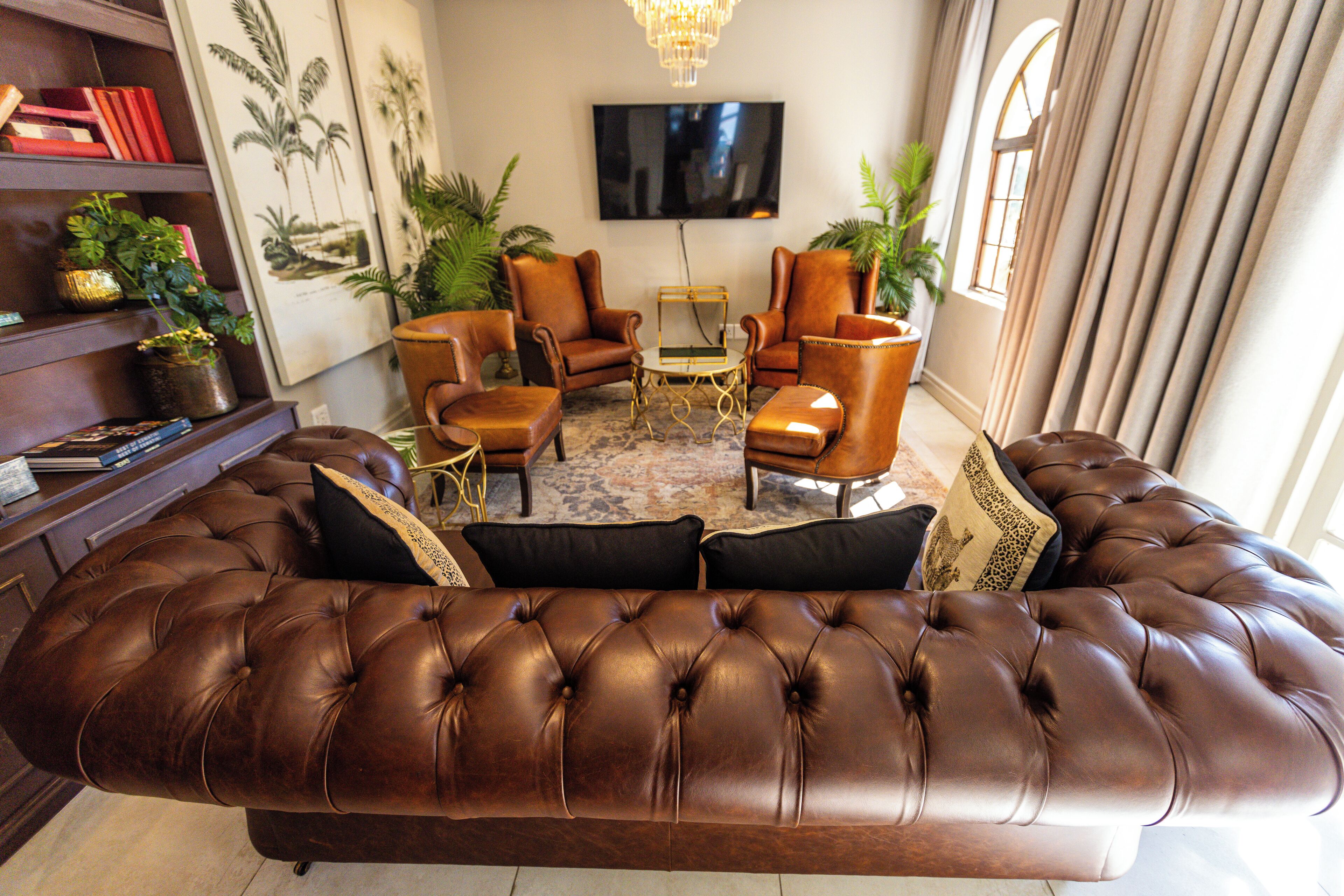 A brown leather sofa with black and patterned cushions, a coffee table, and two armchairs in a well-lit room with a television on the wall and a large window with curtains.