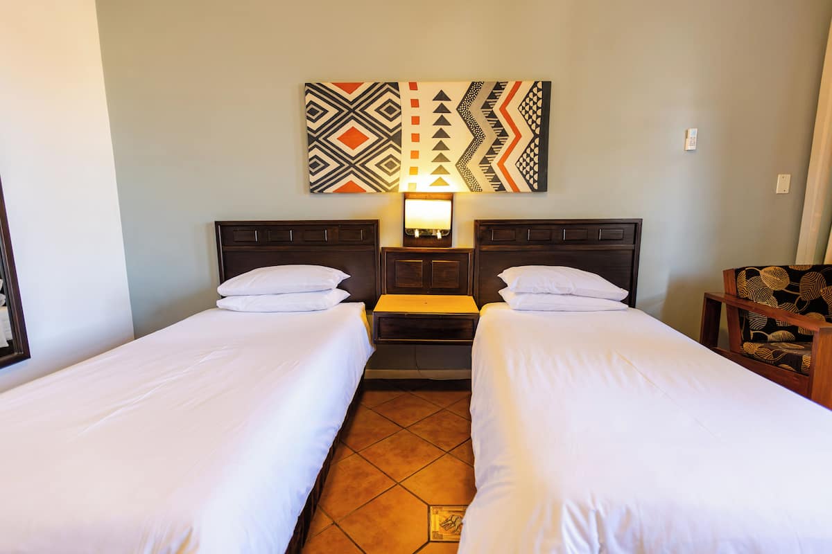 A hotel room with two single beds, a nightstand, a lamp, and a wall-mounted artwork.