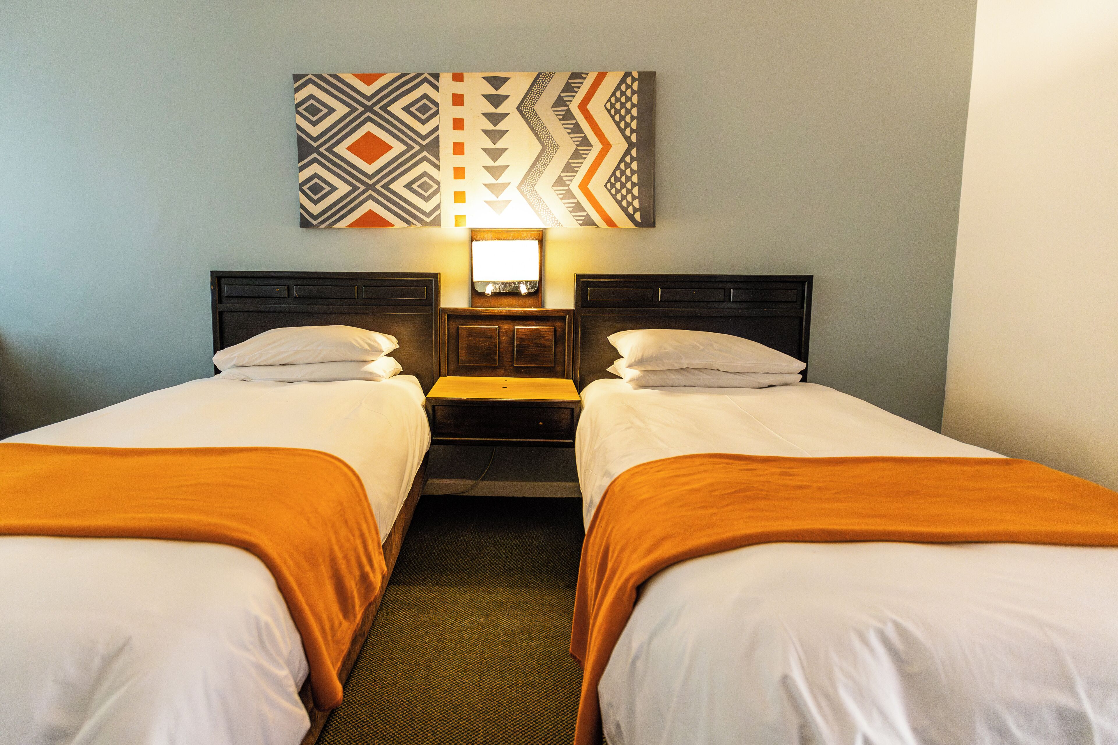 A hotel room with two beds, a nightstand, a wall-mounted TV, and a patterned artwork above the bed.