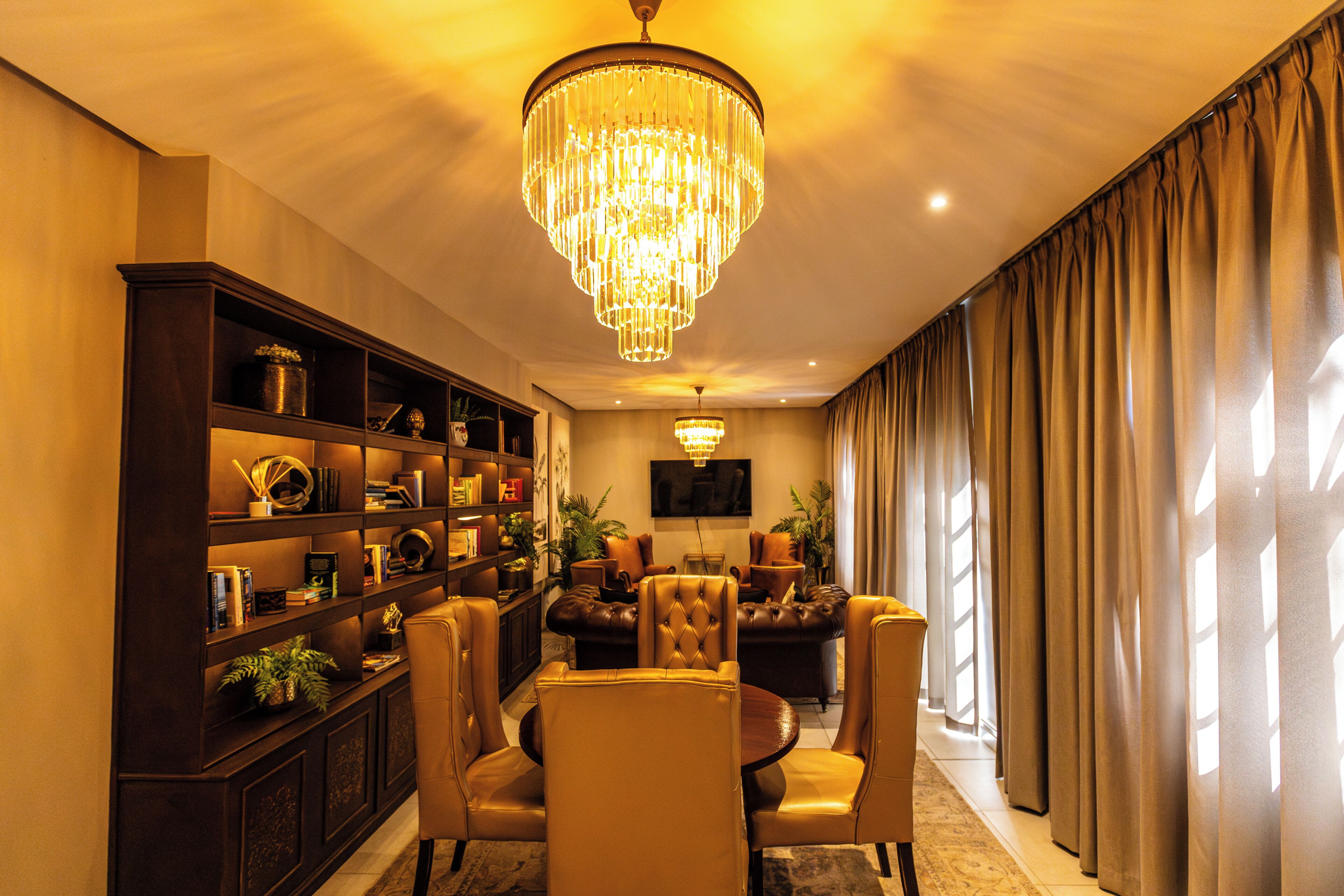 A well-lit living room with a chandelier, leather sofas, a dining table with chairs, a bookshelf, and a television.