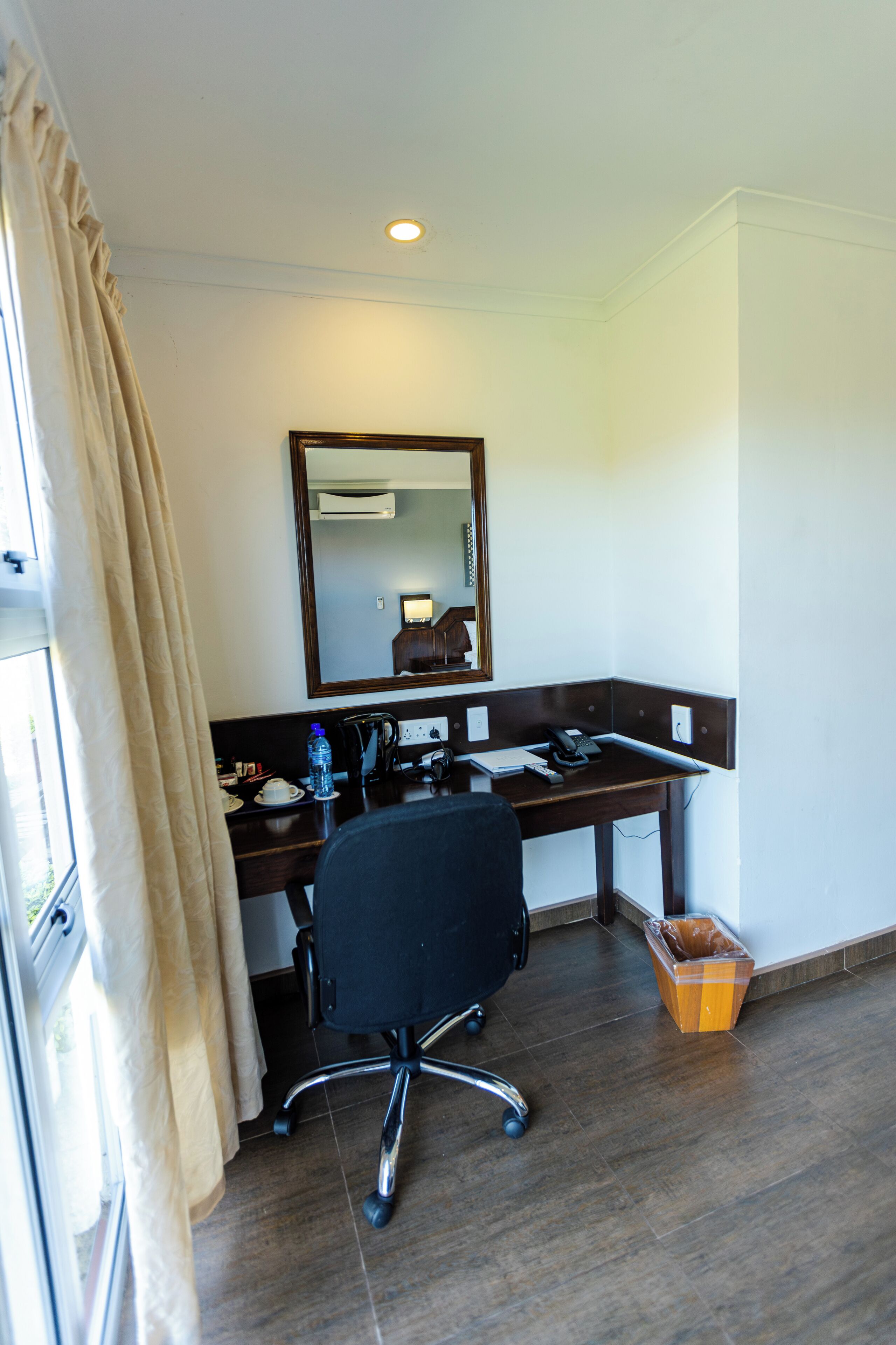 A hotel room with a desk, chair, mirror, and a trash bin.
