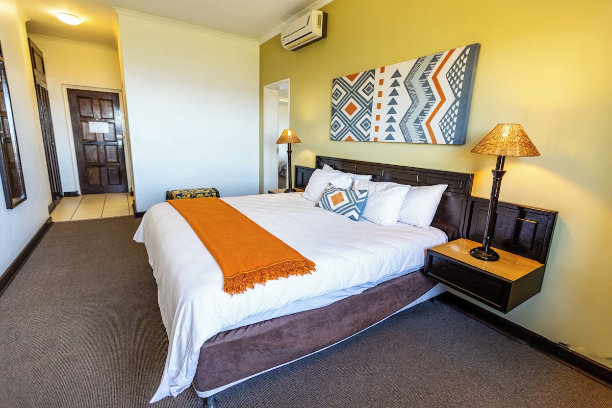 A hotel room with a large bed, a nightstand with a lamp, a wall-mounted air conditioner, and a door leading to another room.