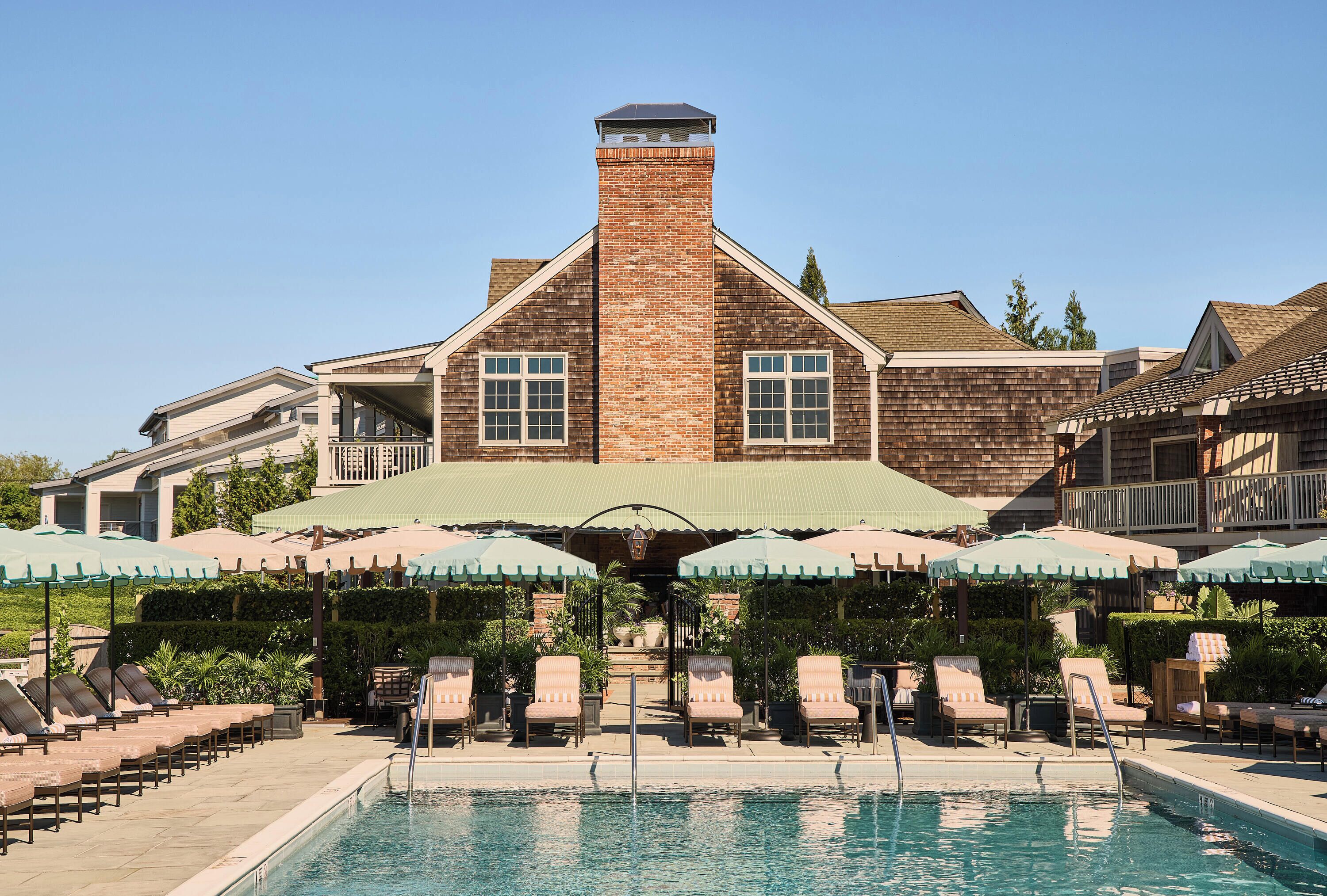 A pool area with lounge chairs and umbrellas, a brick building with a chimney, and a clear blue sky.