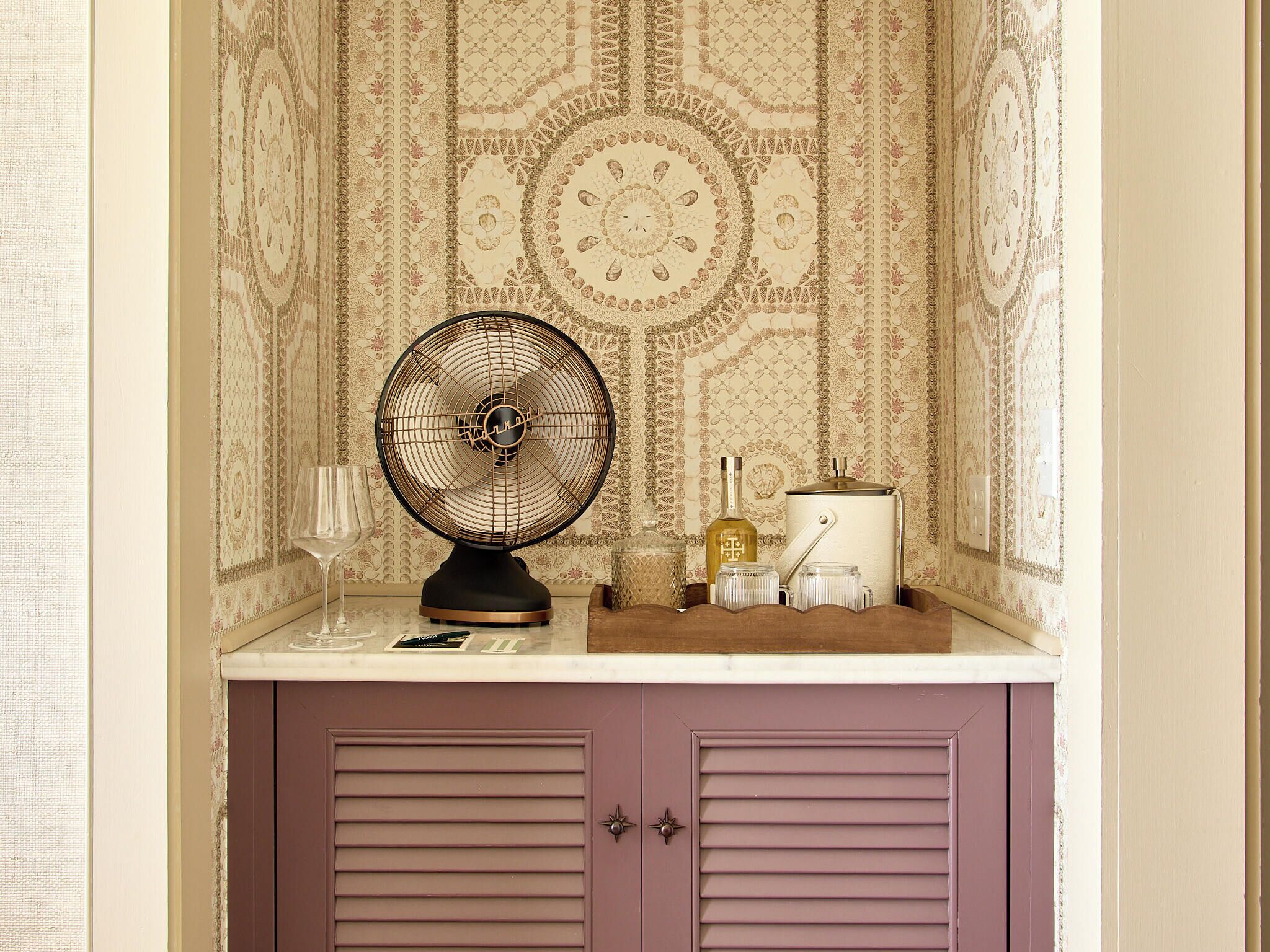 Un ventilateur ancien, une bouteille et un petit récipient sont posés sur un comptoir, devant un papier peint à motifs et une porte violette.