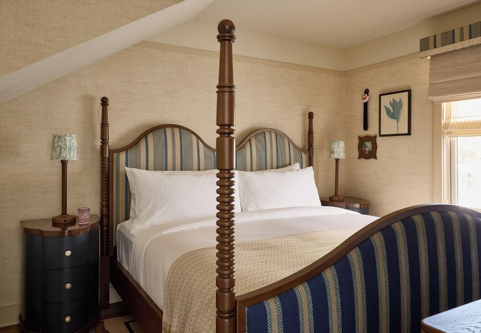 A four-poster bed with a wooden frame and upholstered headboard, a bedside table with a lamp, and a wall-mounted picture frame.