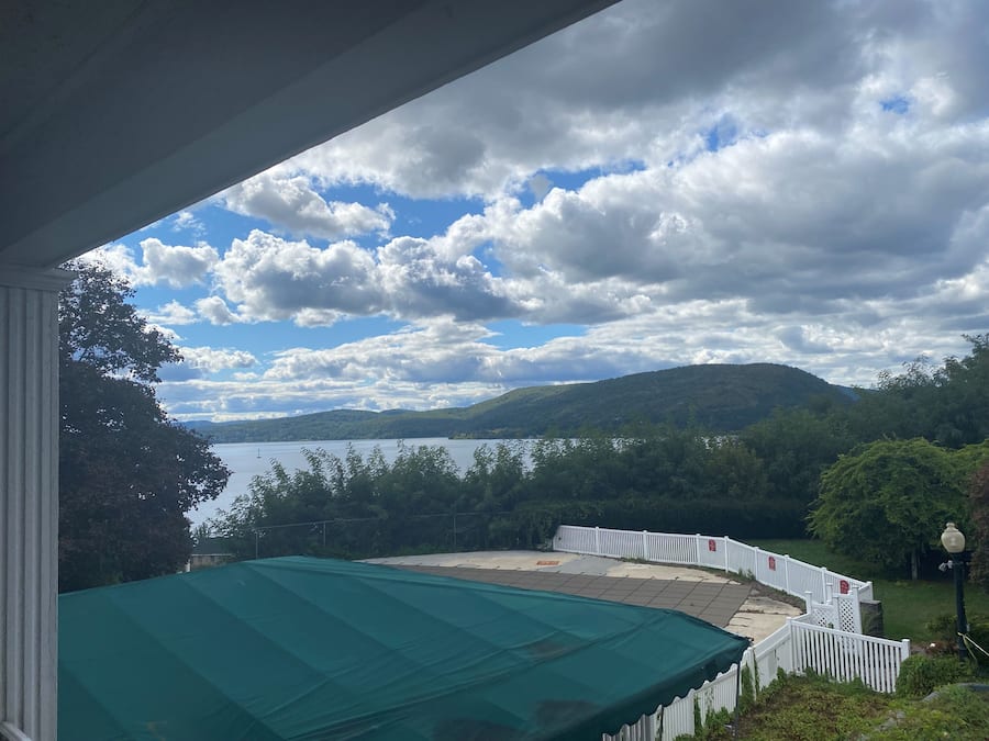 A view from a covered porch overlooking a lake, with a white fence and trees in the foreground.