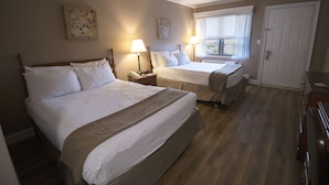 Une chambre d’hôtel avec deux lits, une grande fenêtre avec des rideaux, une porte et une petite table avec une lampe.