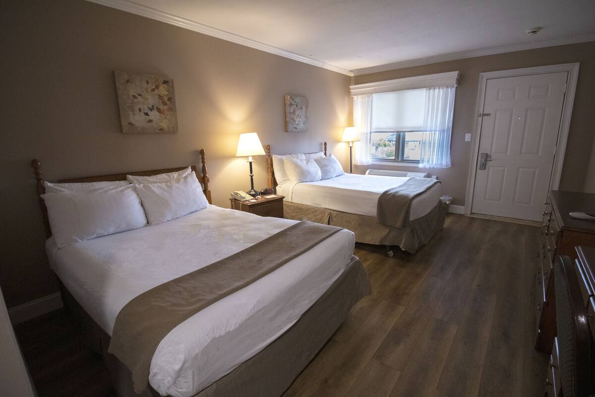 Habitación de hotel con dos camas, un ventanal grande con cortinas, una puerta y una mesita con lámpara.
