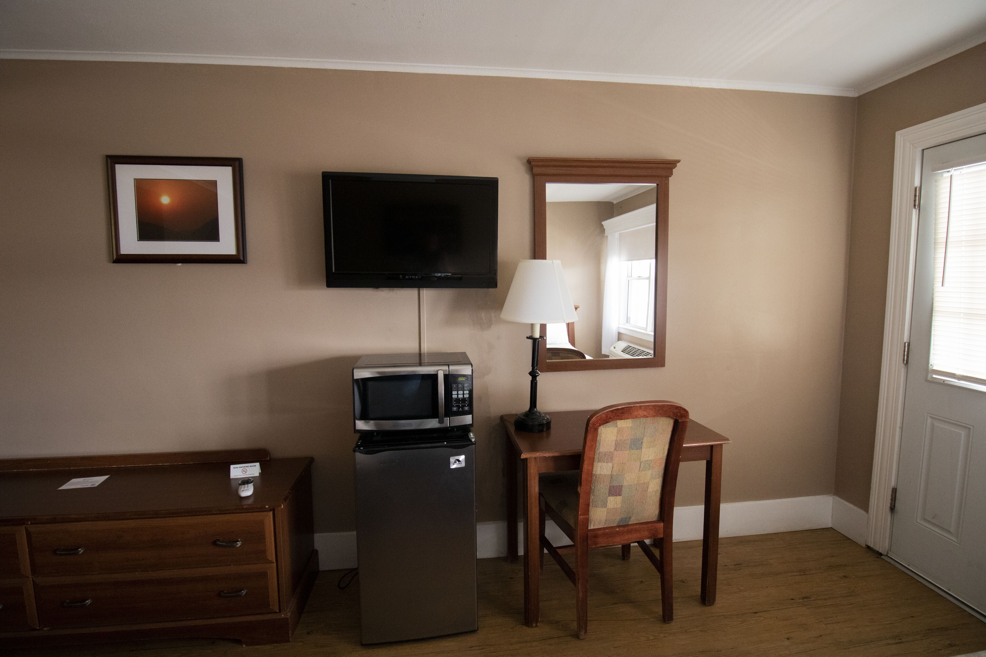 A compact room with a wooden desk, a chair, a television mounted on the wall, a microwave, and a refrigerator.